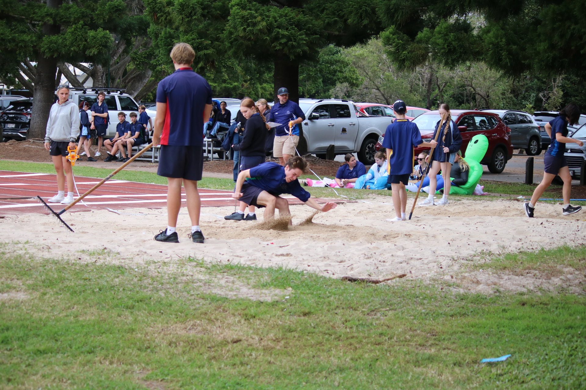 Students participating in a long jump event with others standing nearby on the field.
