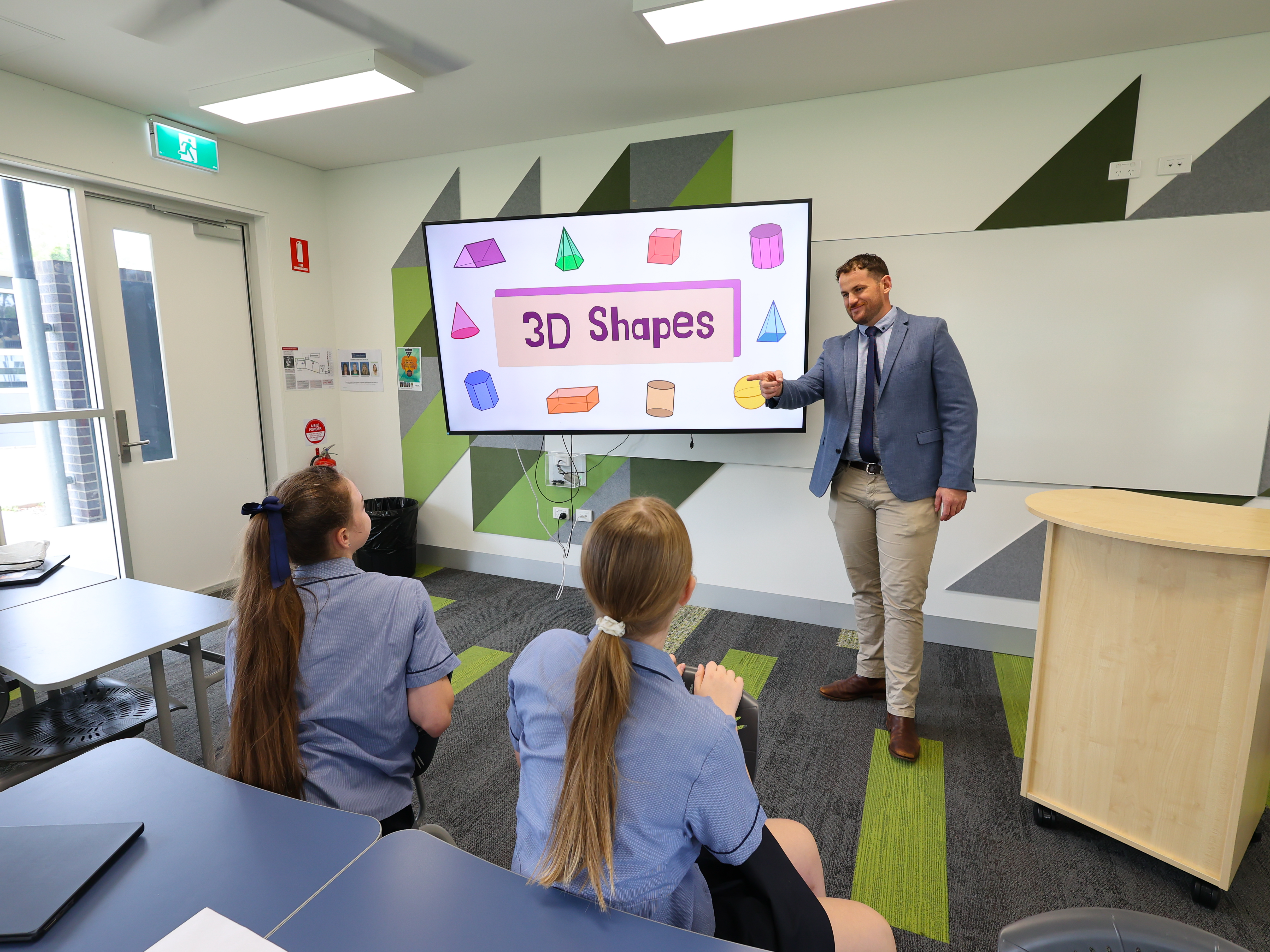 Classroom scene with a teacher showing 3D shapes on a screen to two students